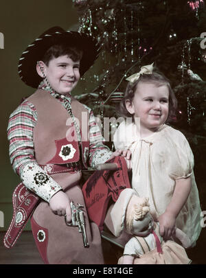 1950 SMILING BOY habillé en tenue de COWBOY HAT CAP HOLDING PISTOL LITTLE GIRL HOLDING A DOLL BY CHRISTMAS TREE Banque D'Images