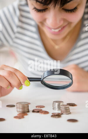 Mixed Race woman examining des piles de pièces de monnaie avec loupe Banque D'Images