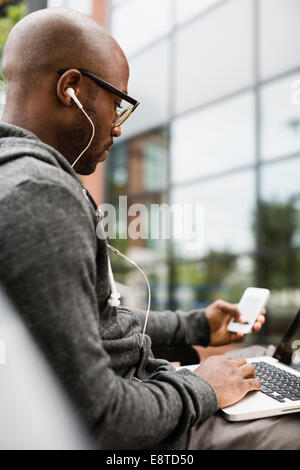 Black man using laptop, écouteurs et cell phone outdoors Banque D'Images