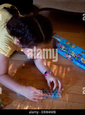 Une mignonne petite fille tisse un bracelet à l'aide d'un arc-en-ciel, un jouet populaire utilisé pour tisser des bandes de caoutchouc en bracelets et charms. Banque D'Images