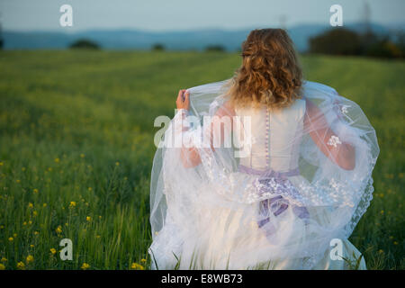 Le Runaway bride - une jeune femme fille dans une robe de mariage qui se tient debout dans un champ de fleurs jaunes soir Banque D'Images