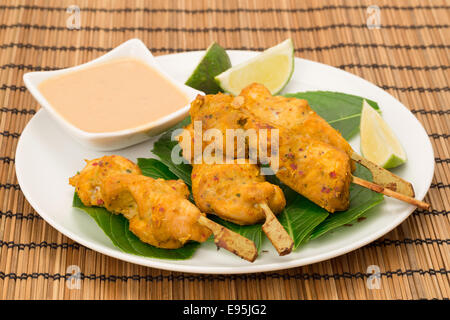 Brochettes de poulet satay servi sur des feuilles vertes avec des tranches de citron vert et d'une trempette aux arachides Banque D'Images