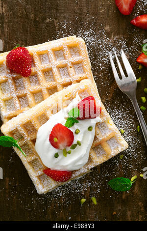 Délicieux gaufre garnie de crème fouettée et de fruits rouges fraises fraîches saupoudré de sucre et servi sur une surface en bois rustique Banque D'Images