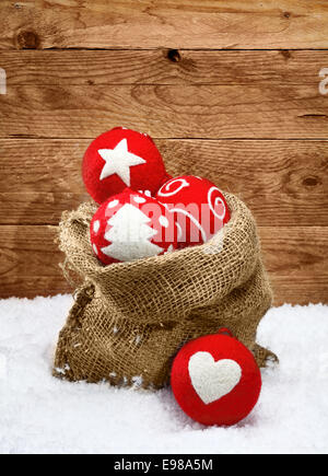 Belle handcrafted rouge décorations de Noël avec des formes traditionnelles décrites sur les boules de tissu tomber d'un sac en toile de jute sur la neige fraîche avec un fond de bois Banque D'Images