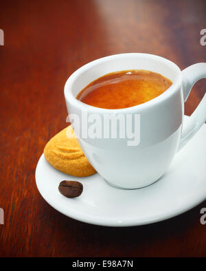 Tasse de café espresso mousseux fraîchement préparé servi avec un biscuit doré et seul grain de café sur une table en bois sombre. Banque D'Images