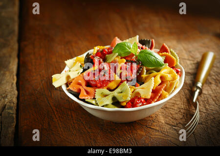 Noeud papillon italien multicolores surmontées de pâtes tomate et garnir de basilic frais servi dans un bol en céramique blanche avec une fourche sur une vieille table en bois Banque D'Images