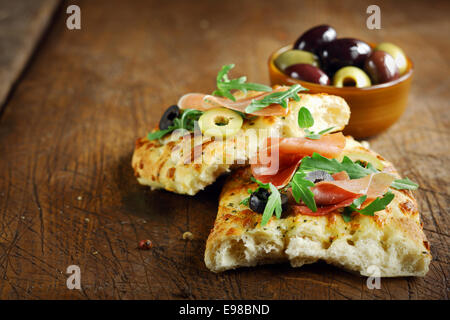 Jambon et à la roquette sur pain focaccia italienne fraîche garnie d'olives tranchées allongé sur une vieille table de cuisine en bois haut Banque D'Images