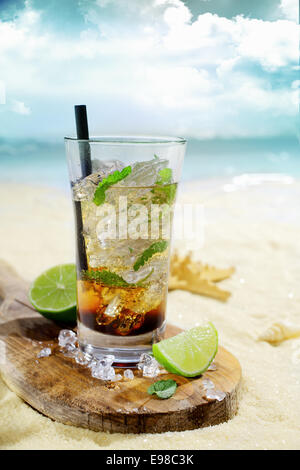 Cocktail au rhum glacé rafraîchissant servi dans un grand verre sur une planche en bois avec des tranches de citron sur le sable doré d'une plage tropicale idyllique Banque D'Images