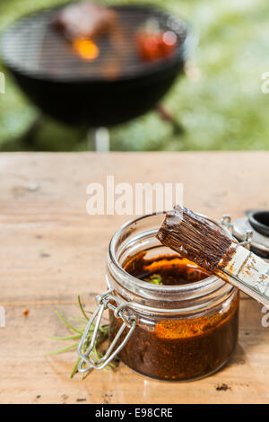 Pot de sauce et un pinceau debout sur une table de pique-nique en bois à l'extérieur pour les viandes grillées sur le barbecue dans l'arrière-plan sur une pelouse verte Banque D'Images