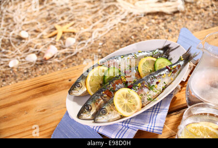 Trois sardines grillées sur un plat sur une table en plein air sur la plage avec un filet de pêche et une étoile de mer en arrière-plan Banque D'Images