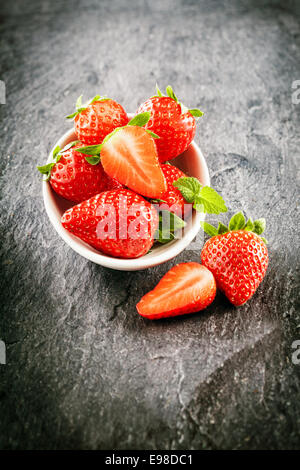 Frais de la ferme de fraises juteuses délicieux servi dans un bol avec l'ensemble et réduit de moitié les fruits dans un high angle view sur ardoise foncé avec vignettage Banque D'Images