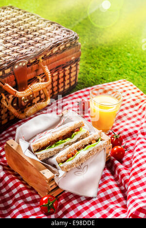 Pique-nique d'été frais, sandwiches tomates cerise et un verre de jus d'orange rafraîchissant sur un tissu à carreaux blancs et rouges avec un panier on Green grass Banque D'Images