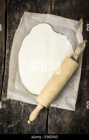 Mis en place une base de pâte à pizza vu de passage lors de la préparation d'une délicieuse pizza italienne avec un rouleau à pâtisserie en bois au-dessus d'elle sur une table en bois rustique grungy Banque D'Images