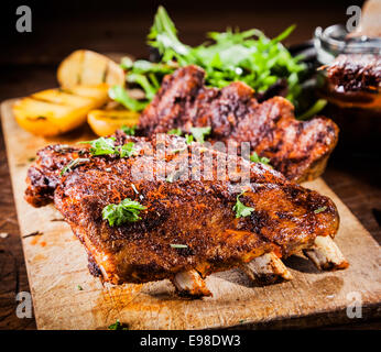 L'arôme délicieux des côtes levées cuites sur un barbecue d'été et mariné avec une sauce épicée garni de fines herbes et servi avec des pommes de terre grillées sur une planche de bois Banque D'Images