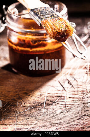 Bocal en verre de sauce épicé fait maison et un pinceau sur une vieille planche en bois marqué dans une cuisine rustique Banque D'Images