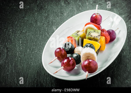 Brochettes de fruits exotiques colorés servis sur un plat ovale avec raisins, kiwis, oranges, bananes, bleuets, fraises et sur un fond sombre avec copyspace Banque D'Images