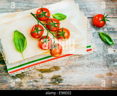 Les tomates cerises, le basilic et les feuilles de lasagne séchée sur une vieille table en bois vieilli dans une cuisine rustique prêts à être utilisés comme ingrédients d'un plat de pâtes italiennes, vue d'en haut Banque D'Images