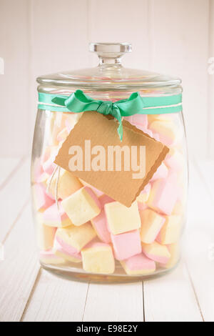 Verre ancien bocal rempli de bonbons carrés avec guimauves rose et jaune et un blanc tag attaché avec un ruban vert sur un fond de bois blanc Banque D'Images