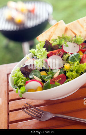 Fruits frais Légumes Légumes verts à salade végétarienne mixtes sur une table de pique-nique en bois avec des tranches de pain grillé et d'un barbecue d'été visibles sur la pelouse au-delà Banque D'Images