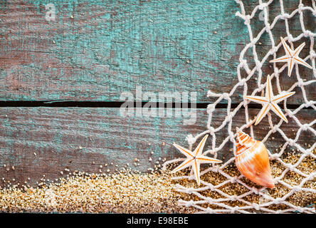 Fond marin rustique avec copyspace sur planches en bois patiné bleu turquoise décorée de filets de pêche à mailles en losanges, étoiles et d'un coquillage sur un lit de sable de plage, des souvenirs de vacances d'été Banque D'Images