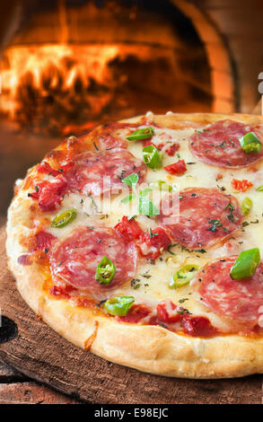 Traditionnels fraîchement préparés ou pepperoni pizza salami italien sur une croûte à tarte en or en face d'un four à pizza au feu dans un restaurant ou pizzeria Banque D'Images