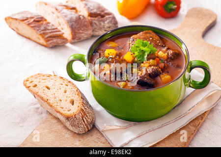 L'hiver savoureux ragoût dans un pot en métal vert avec de la viande et de légumes variés dans une sauce riche servi avec pain croûté frais sur une planche à découper en bois Banque D'Images