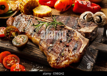 Close up d'un succulent steak d'aloyau grillé tendre assaisonné de poivre et de romarin sur une planche en bois avec des tomates coupées en deux, les champignons, les épis et les poivrons Banque D'Images