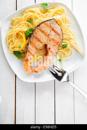 La cuisine de fruits de mer gastronomique avec escalope de saumon grillé steak servi sur un lit de pâtes linguine italienne garnie avec citron et basilic frais, vue aérienne sur une table en bois blanc avec copyspace Banque D'Images