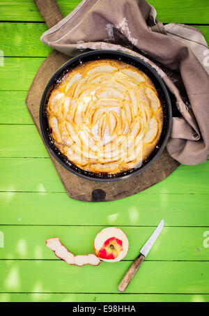 La tarte aux pommes faite maison cuit avec des pommes fraîches forment le jardin servi sur une table de jardin en bois vert tacheté dans la lumière du soleil avec une demi pomme pelée et couteaux aux côtés Banque D'Images