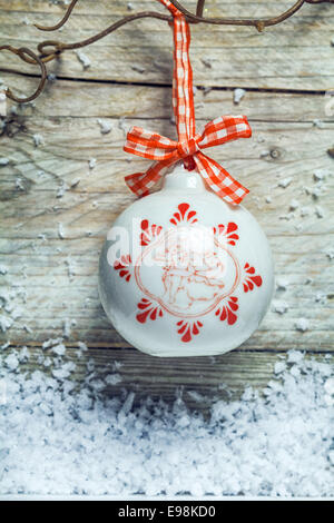 Jolie boule de noël en céramique décorée suspendu par un ruban rouge et blanc vérifié à partir de paires torsadées sur un fond de bois patiné avec neige de l'hiver Banque D'Images