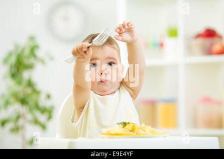 Un an old kid garçon dans une chaise haute pour l'alimentation à l'aide d'une fourchette et Banque D'Images