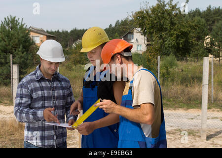 Trois constructeurs dans leurs bâtiments debout dans un groupe de discussion sur la paperasse sur un site de construction, alors qu'ils tentent de résoudre un problème Banque D'Images