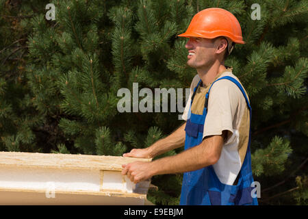 Jeune ouvrier sur un chantier portant un panneau d'isolation des murs en bois avec un sourire, en vue de côté contre verdure avec copyspace. Banque D'Images