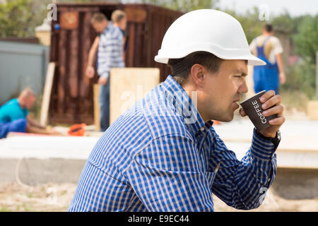 Builder ou construction worker sitting savourer une tasse de café sur place avec ses collègues travaillant dans l'arrière-plan, Vue de côté Banque D'Images