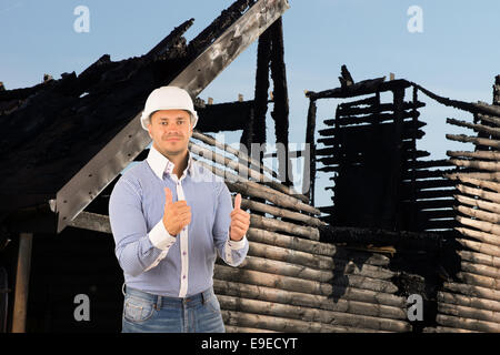 L'âge moyen Ingénieur mâle double montrant Thumbs Up Sign avec le feu maison endommagée. Banque D'Images