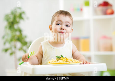 Bébé garçon dans une chaise haute pour l'alimentation à l'aide d'une fourchette et une assiette je Banque D'Images