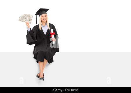 Female graduate holding de l'argent assis sur un panneau isolé sur fond blanc Banque D'Images