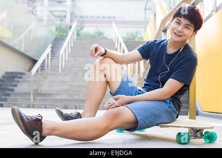 Young man sitting on skateboard listening to earphones Banque D'Images