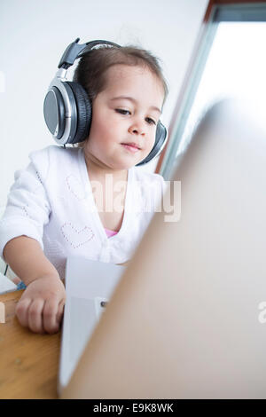 Little girl wearing headphones while looking at laptop at table in house Banque D'Images