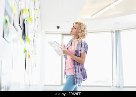 Creative businesswoman looking at papers coincé sur le mur lors de l'écriture notes in office Banque D'Images