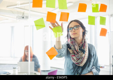 Creative businesswoman reading notes adhésives sur mur de verre avec collègue travaillant en arrière-plan à l'office de tourisme Banque D'Images