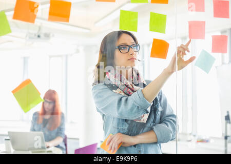 Creative businesswoman reading notes adhésives sur mur de verre avec collègue travaillant en arrière-plan à l'office de tourisme Banque D'Images