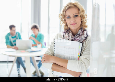 Portrait of businesswoman holding Création de fichiers avec des collègues qui travaillent en arrière-plan à l'office de tourisme Banque D'Images