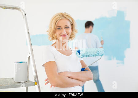 Portrait of woman holding paint roller avec man painting wall en arrière-plan Banque D'Images