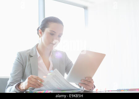 Young businesswoman using tablet PC while reading book in office Banque D'Images