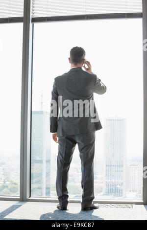 Vue arrière sur toute la longueur de mature businessman using cell phone white debout près de window Banque D'Images