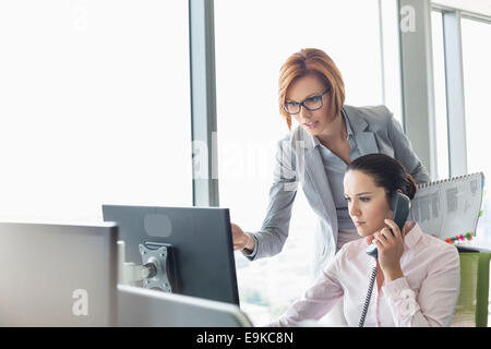 Young businesswoman using téléphone fixe le collègue tout en pointant sur le moniteur de l'ordinateur de bureau Banque D'Images