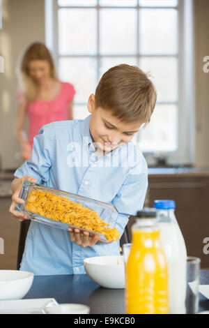 Boy pouring flocons de maïs dans un bol avec la mère debout en arrière-plan Banque D'Images