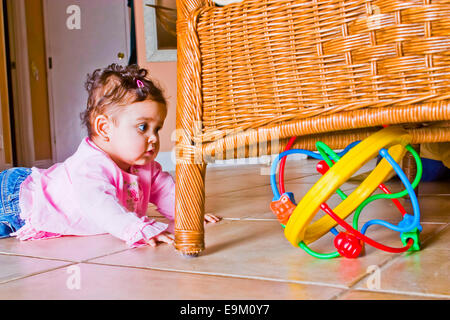 9-month-old baby girl colombien se traîne vers toy de couleur vive caché sous le rotin fauteuil à la maison. M. © Myrleen Pearson Banque D'Images