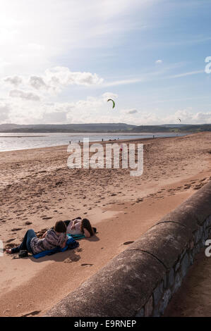 Deux étudiantes vous allonger sur la plage à Exmouth en regardant vers l'estuaire de la rivière Exe, regarder des kite surfeurs. Banque D'Images
